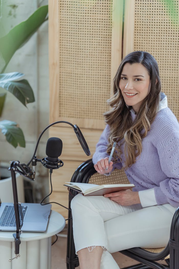 who-we-are Young positive lady in casual clothes sitting in armchair near laptop and microphone with notepad on knees near cabinet and potted plant while looking at camera in light room